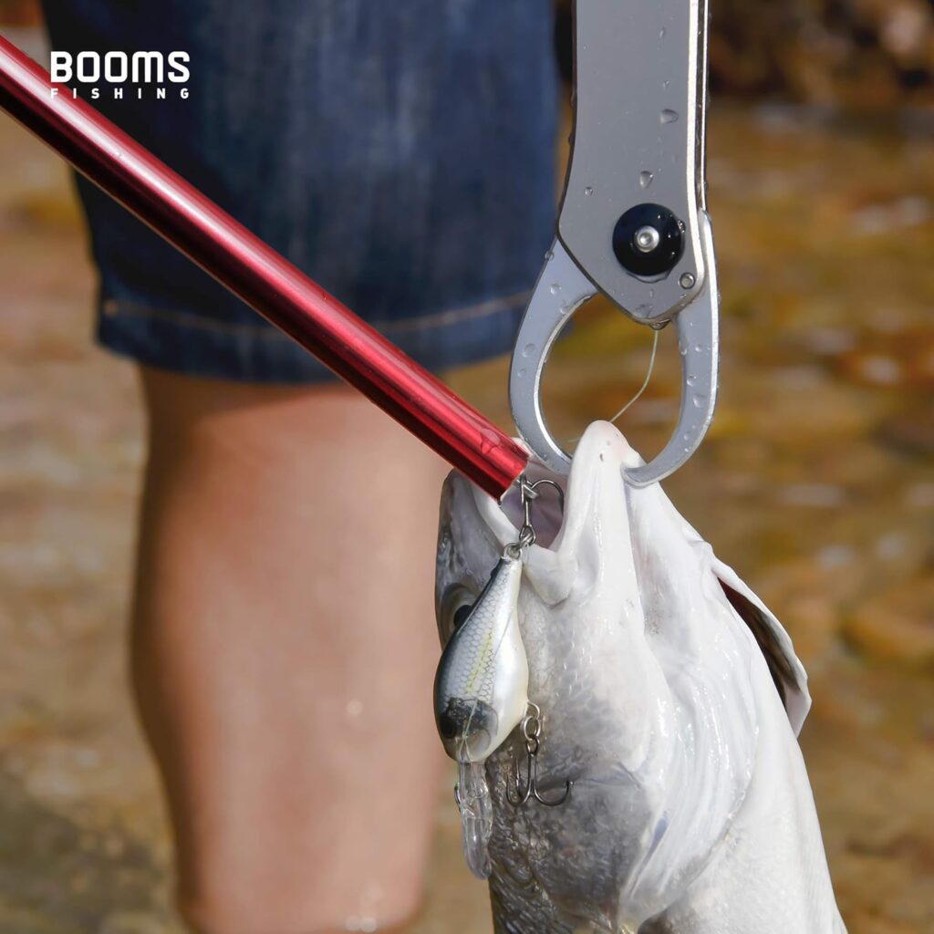 A close-up of a fish held by fishing pliers, with a red fishing rod and lure in its mouth. Perfect for Fishing Gifts or Cool Gifts for Men, while someone stands in shallow water. The text BOOMS FISHING appears in the top left corner. gift idea