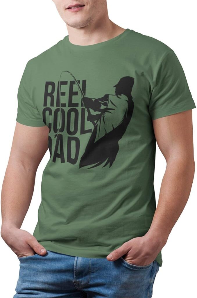 A man wearing a green T-shirt with a black silhouette of a person fishing and the text REEL COOL DAD printed on the front—an ideal pick for Fishing Gifts. He has his hands in his pockets and is wearing blue jeans. gift idea
