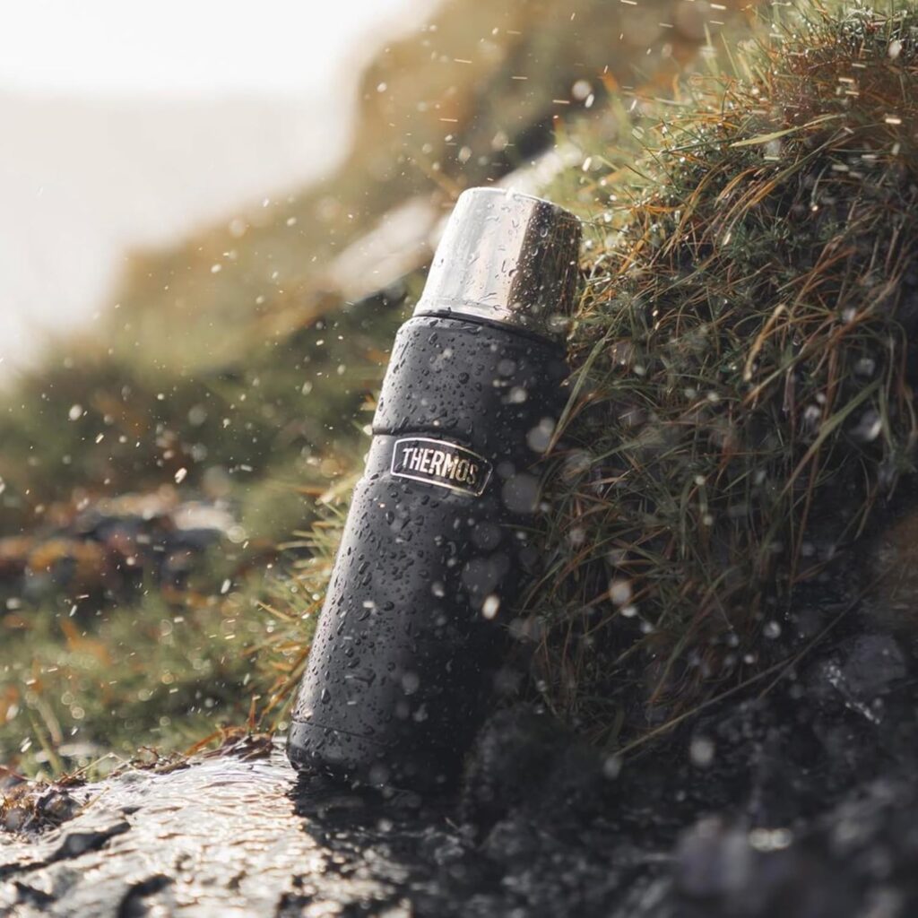 A black Thermos flask with a silver lid sits on wet grass and moss, covered in water droplets, with rainfall and blurred natural scenery in the background—perfect as one of those unique fishing gifts for men who love fishing. gift idea