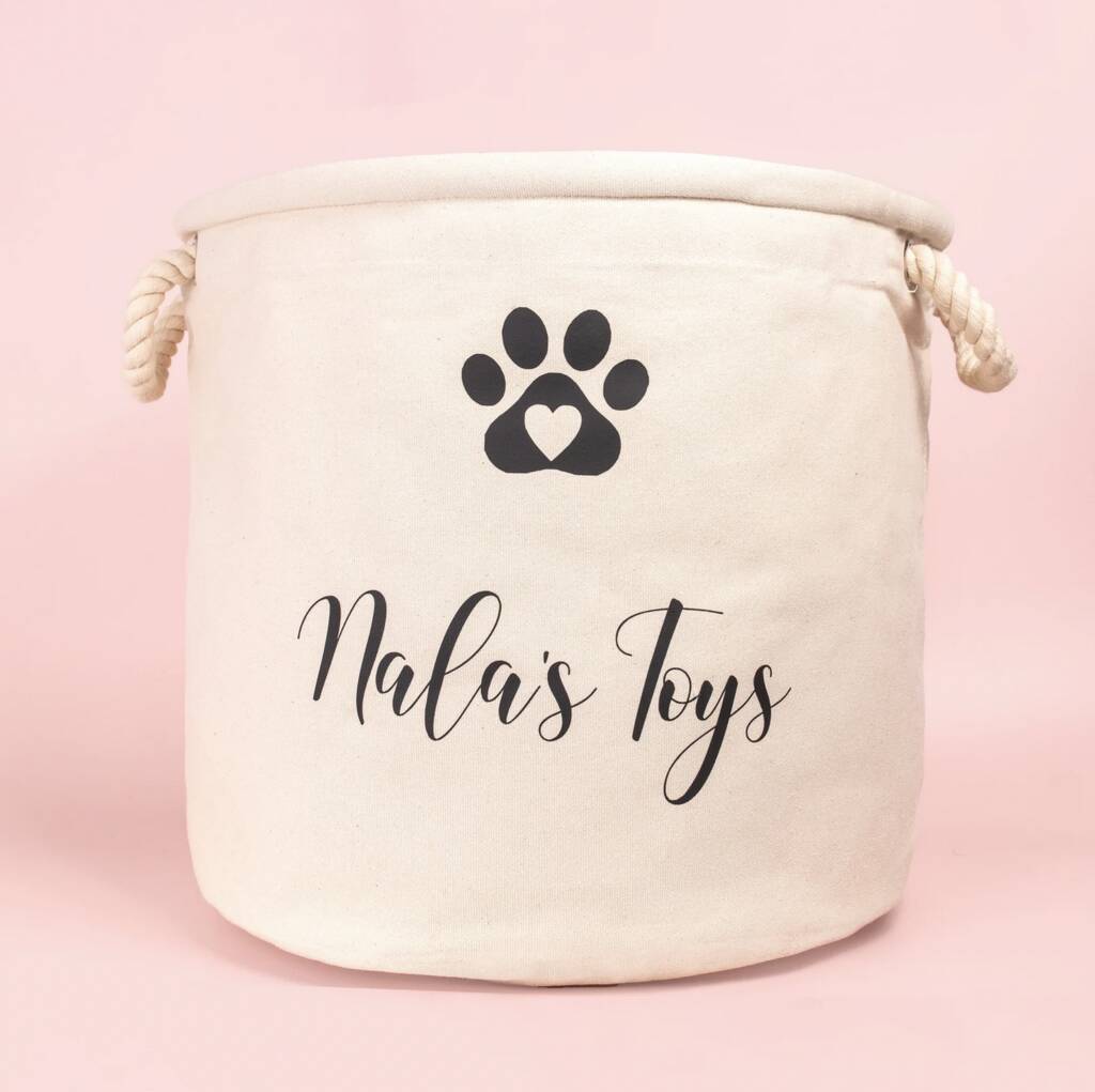 A cream-colored fabric storage bin with rope handles, featuring a black paw print with a heart and the words Nalas Toys in stylish script on the front—a perfect gift idea for any pet lover, set against a light pink background. alwaysagift.co.uk