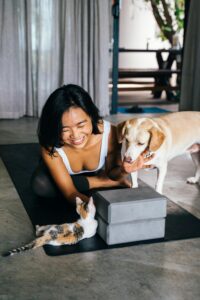 A woman smiles while kneeling on a yoga mat indoors, reaching toward a cat lying near two stacked yoga blocks, as a dog stands nearby—capturing a sweet moment thats also a perfect gift idea for pet lovers. alwaysagift.co.uk