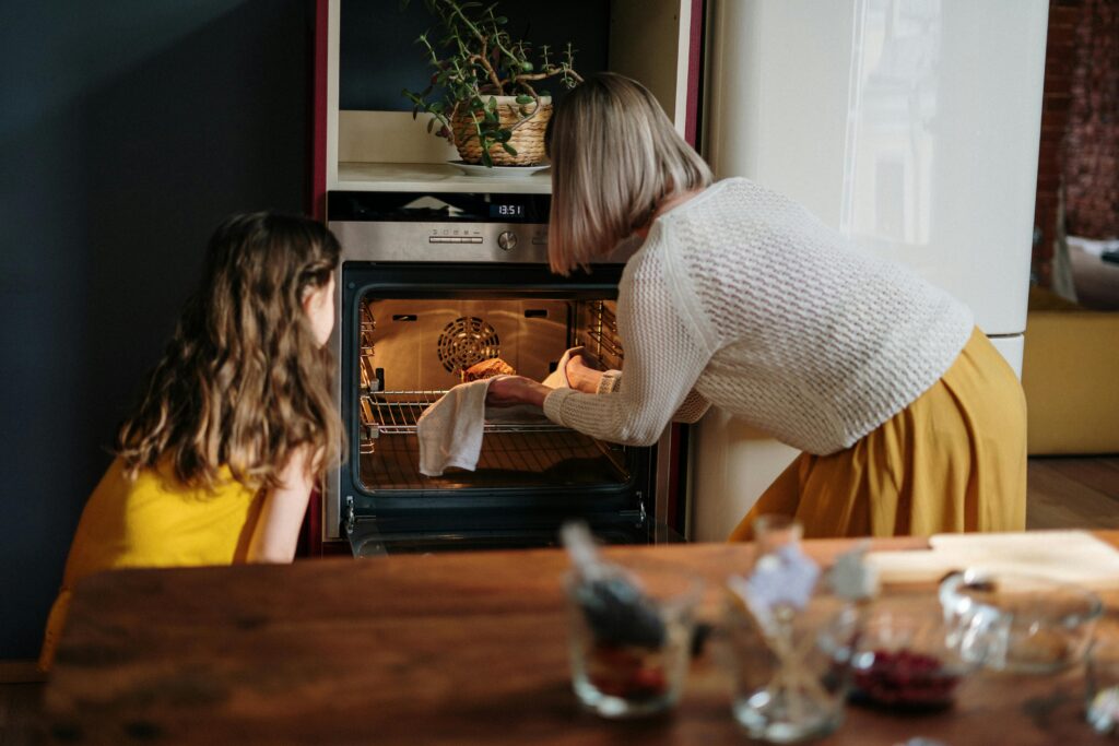 A woman and a young girl are looking into an open oven in a kitchen, perhaps checking a freshly baked treat—perfect inspiration for gift ideas for women or thoughtful baking gifts. A plant sits on the oven and jars rest on the table in the foreground. gift idea
