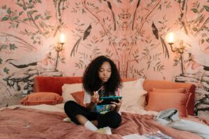 A girl with curly hair sits cross-legged on a pink bed, holding a tablet and stylus—making this cozy setup a perfect gift idea. The room features pink walls with bird and tree murals, warm wall lamp lighting, pillows, and a jacket on the bed. alwaysagift.co.uk