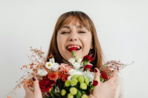 A smiling woman with red lipstick holds a colorful bouquet of fresh flowers—red, white, pink, and green blooms—against a plain white background, making it a perfect gift idea. alwaysagift.co.uk