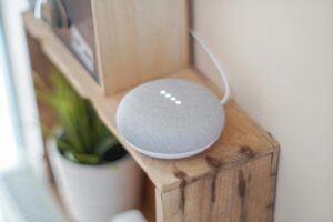 A small, gray smart speaker with four white LED lights sits on a wooden shelf next to a potted plant. A white cord is plugged into the device, adding a modern touch to any Elementor-inspired workspace. alwaysagift.co.uk