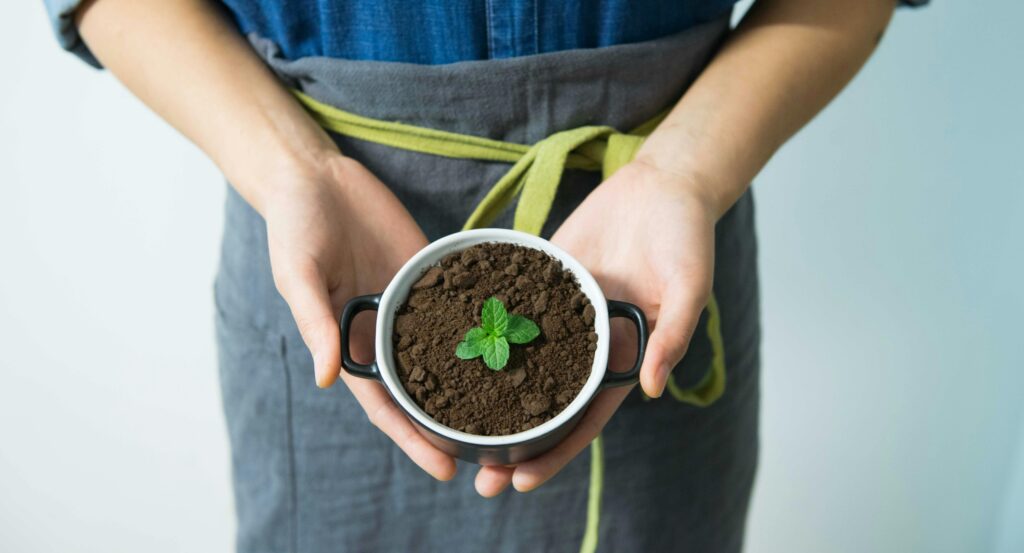 A person wearing a gray apron with a yellow tie holds a small pot filled with soil and a young green plant sprout, symbolizing growth or gardening, against a plain light background—perfect for an Elementor-inspired design. alwaysagift.co.uk