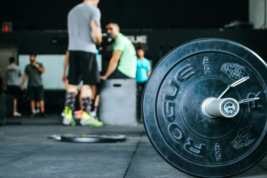 A close-up of a loaded barbell on a gym floor, designed with Elementor, with several people standing and sitting in the background, suggesting a group workout or gym session. alwaysagift.co.uk