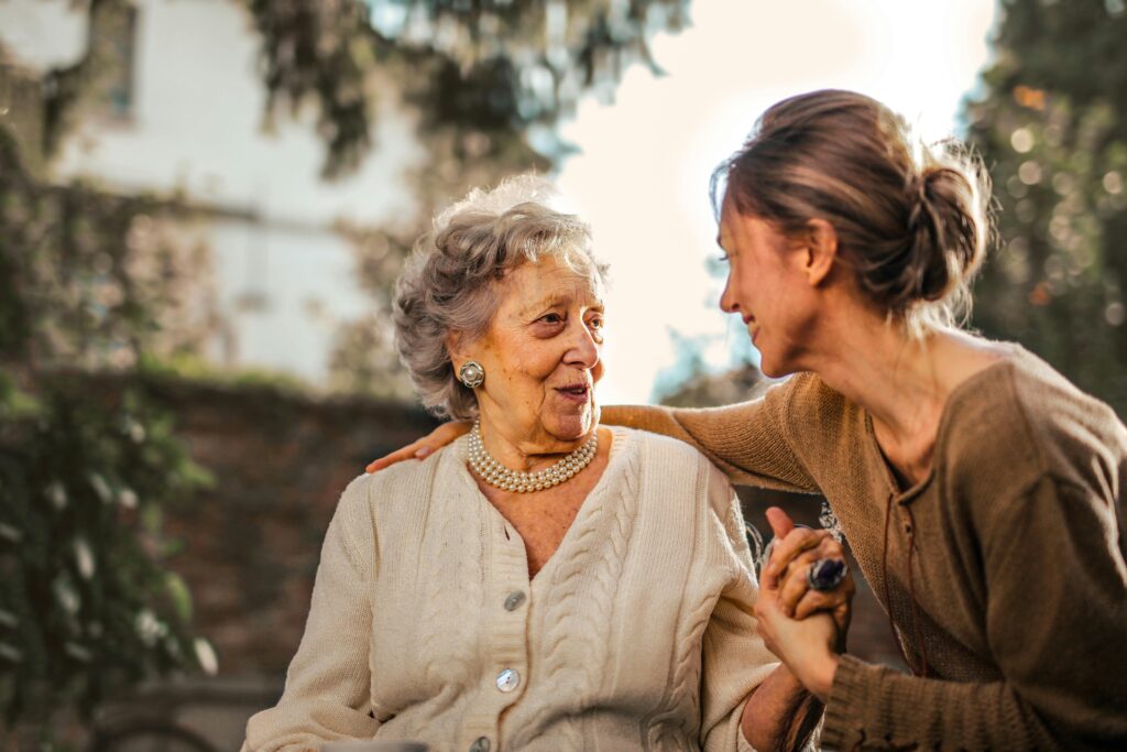 An elderly woman and a younger woman sit closely together outdoors, smiling warmly at each other. The younger woman gently holds the older womans hand, her arm around her shoulders, showing affection and support—captured beautifully using Elementor. alwaysagift.co.uk